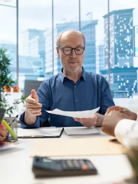 Older man in glasses discussing tax documents at a table with a calculator and various hands visible, emphasizing tax planning and financial consultation.