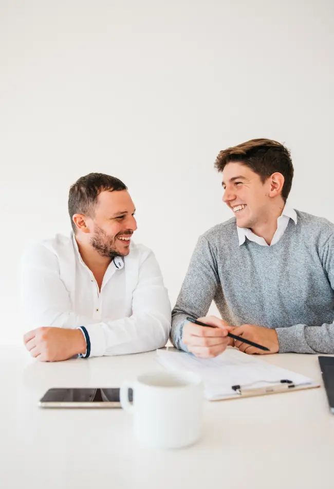 Two men smiling and engaging in conversation at a desk, discussing tax filing services, with a notebook, pen, and coffee cup visible.